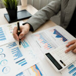 Close-up of a business professional analyzing graphs and charts with a calculator and pen at a desk, representing budget planning and financial strategy.