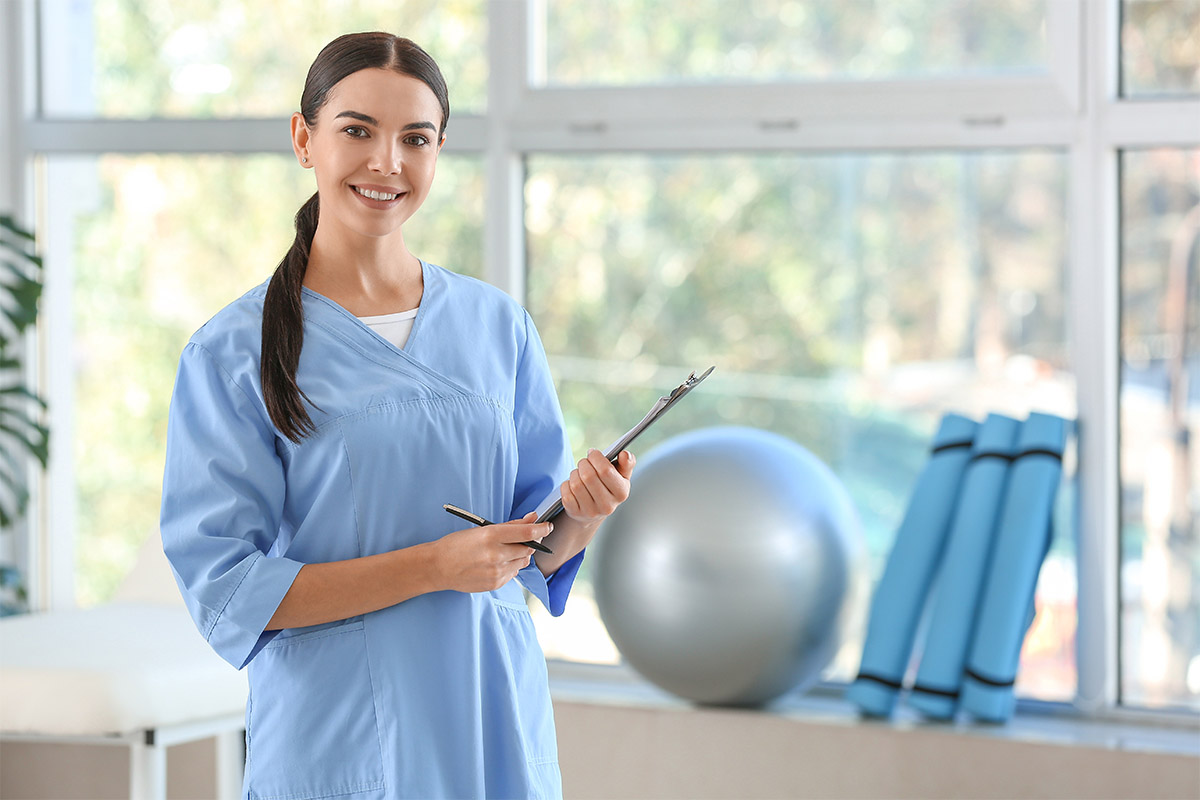 Smiling physical therapist in blue scrubs holding a clipboard in a modern clinic setting with exercise equipment in the background.