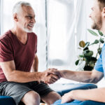 A smiling elderly man in a red t-shirt shakes hands with a healthcare professional in blue scrubs, symbolizing trust and patient loyalty in a medical setting.