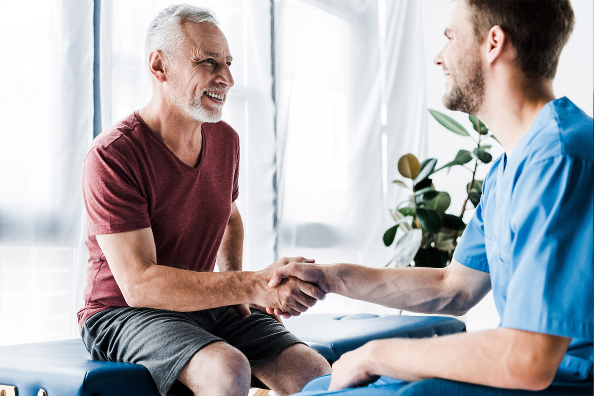 A smiling elderly man in a red t-shirt shakes hands with a healthcare professional in blue scrubs, symbolizing trust and patient loyalty in a medical setting.